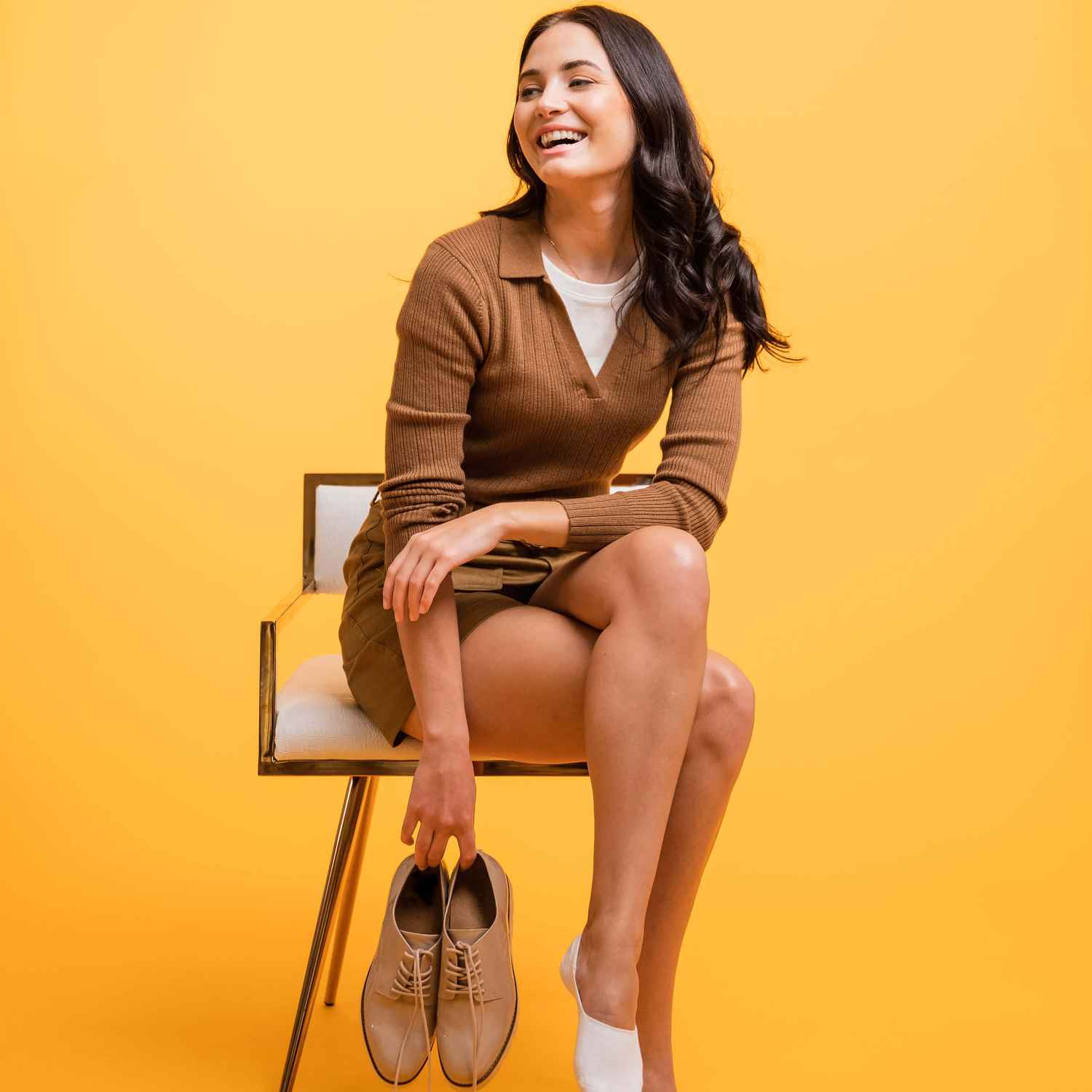 Une femme rit en étant assise sur une chaise et en tenant ses chaussures.