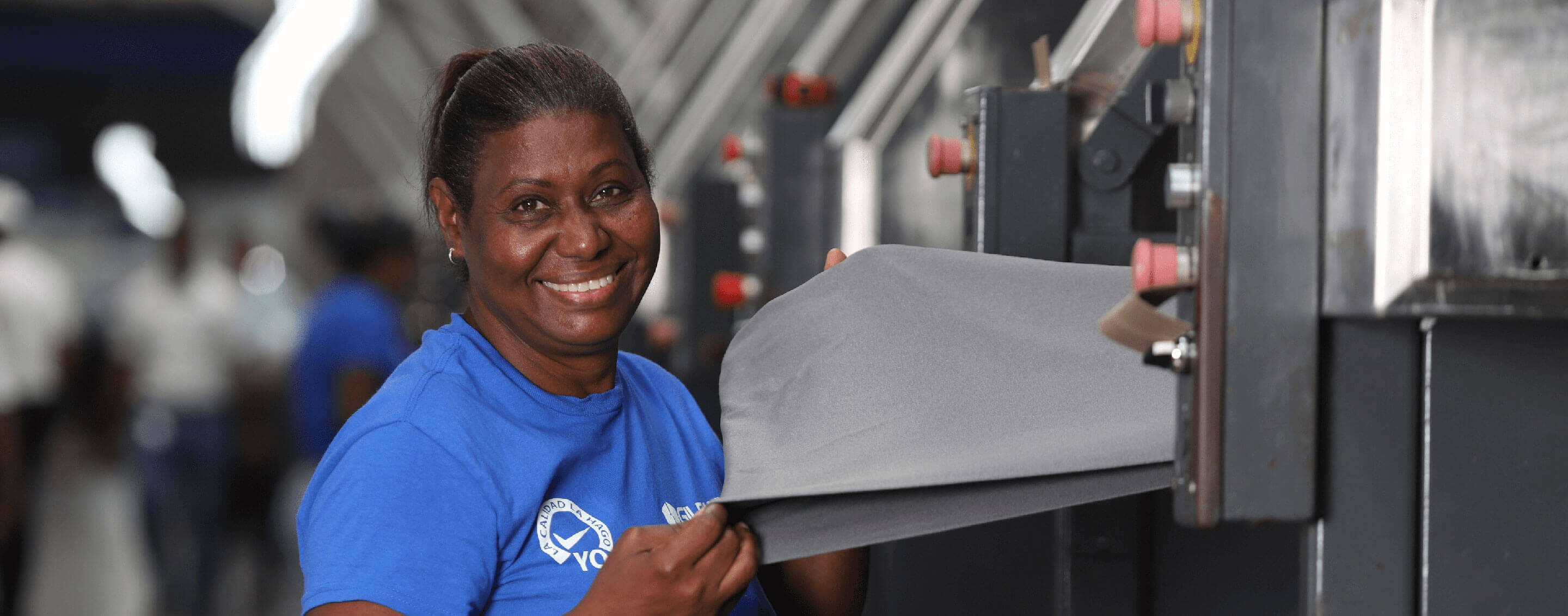 Une employée portant une chemise bleue Gildan tient un morceau de tissu et sourit.