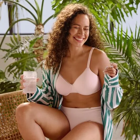 A woman drinking water outside laughing and wearing light pink undergarments with a striped-green shirt.