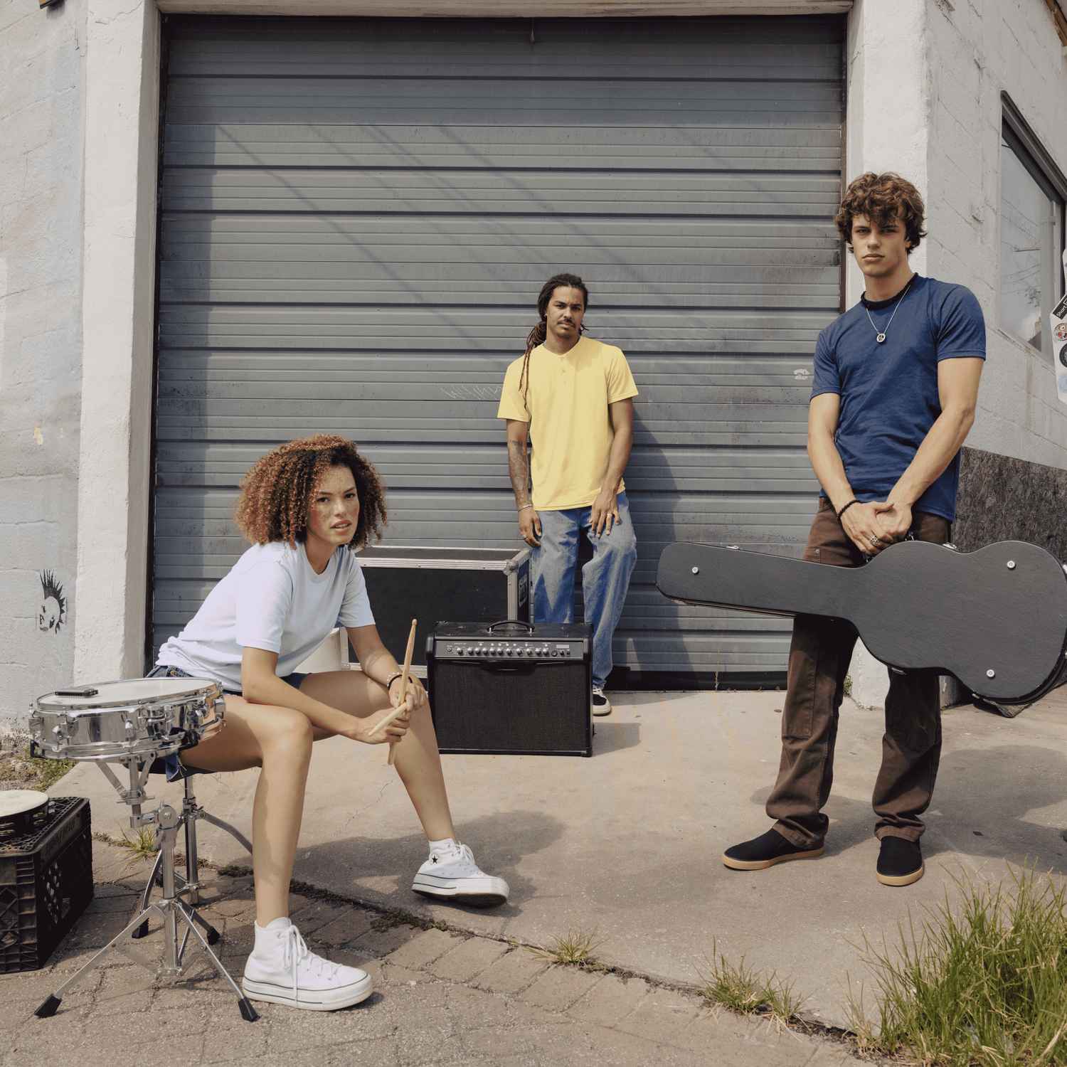 Two men and one woman are outside in front of a garage with band instruments.