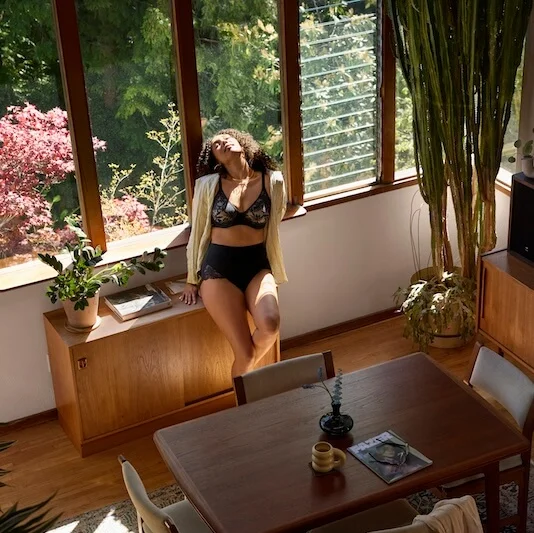 A woman in black innerwear sits by a sunlit window surrounded by plants.