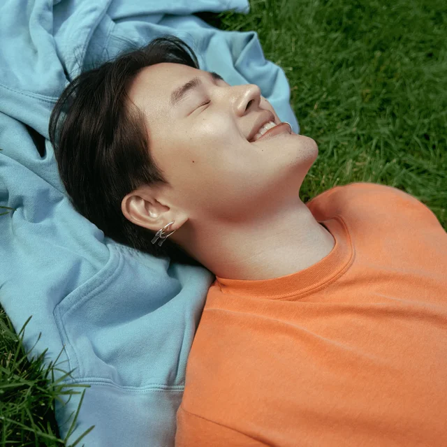 A person wearing an orange Comfort Colors shirt on the grass closing their eyes and smiling at the sun.