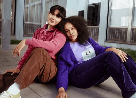 A woman wearing purple and a man wearing pink are sitting on the floor leaning against each other.