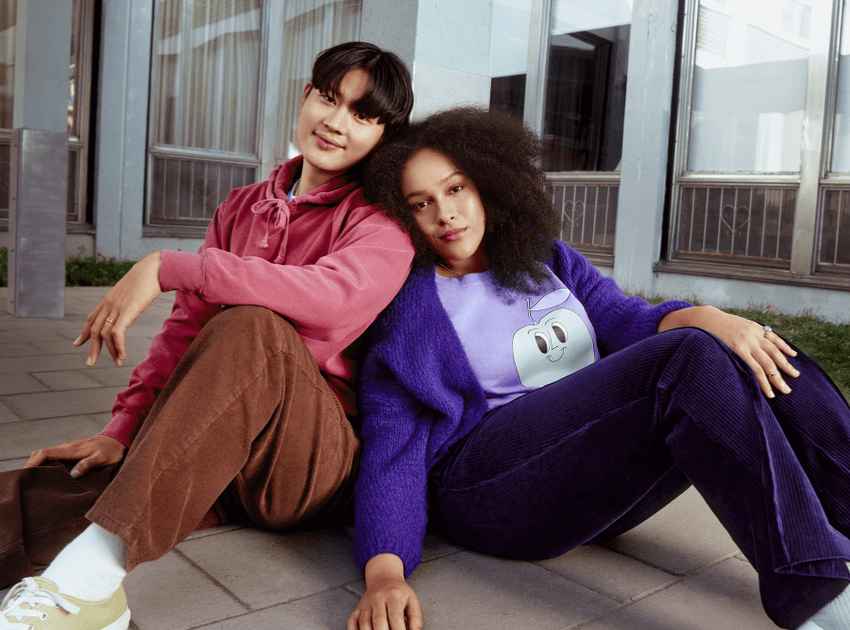 A woman wearing purple and a man wearing pink are sitting on the floor leaning against each other.