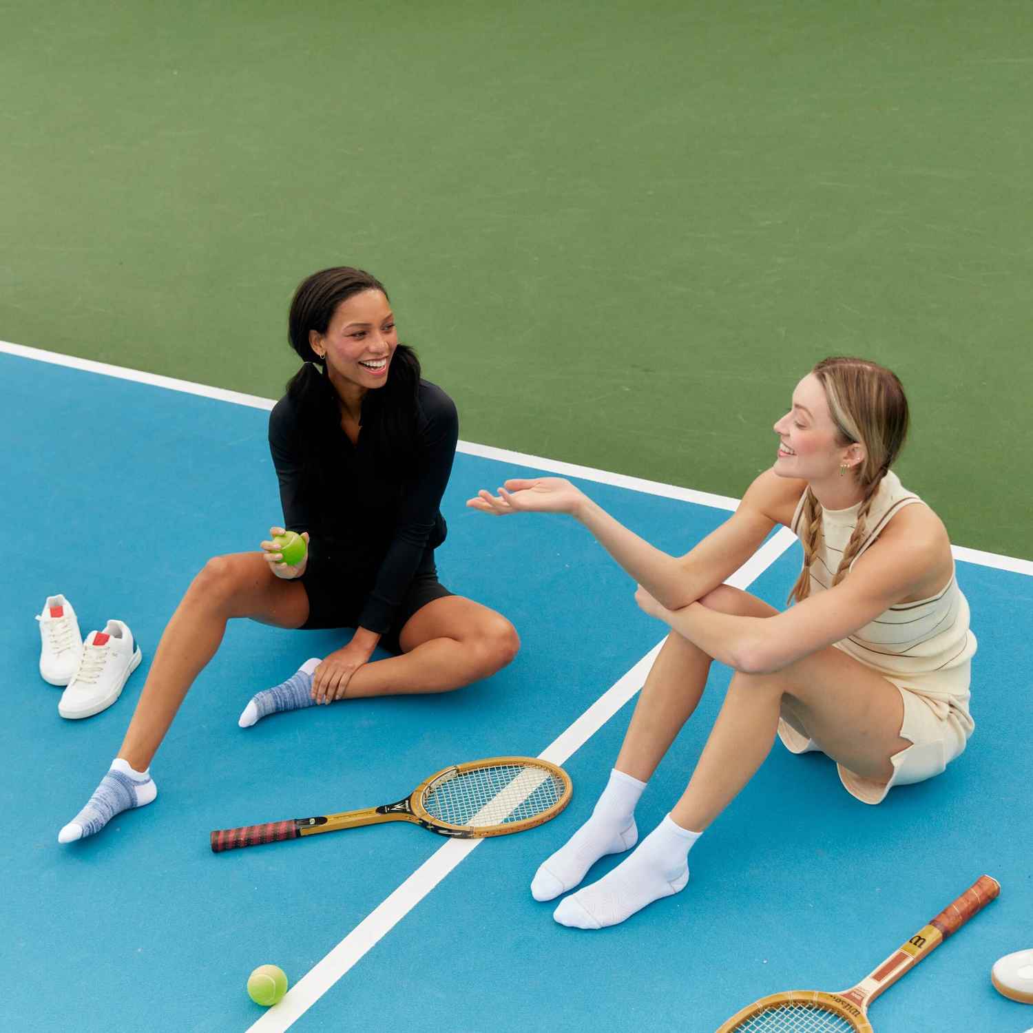 Two women are sitting on a tennis court in their Peds socks talking and smiling.