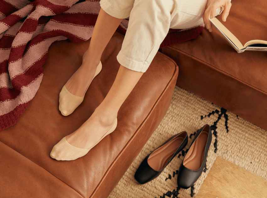 A closeup of a woman's feet wearing Peds no show beige socks on a brown leather couch.