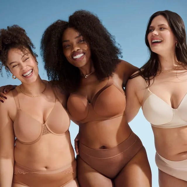 3 women are wearing beige, brown, and white intimates, standing together in front of a blue background.