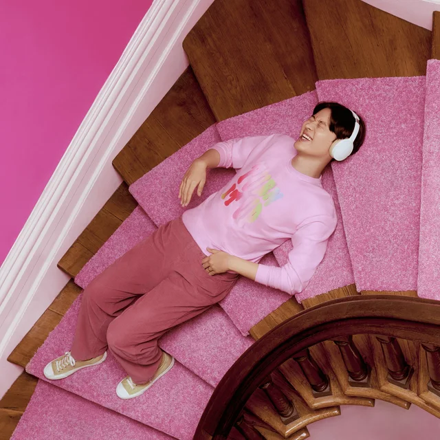 A person wearing a pink Comfort Colors shirt lying down on a staircase wearing white headphones and laughing.