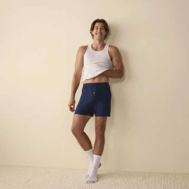 A man leaning against a wall wearing white tank top and navy shorts.