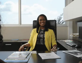 A woman in a yellow suit is sitting in a corporate office and smiling.