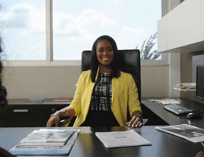 Une femme en tailleur jaune est assise dans un bureau d'entreprise et sourit.