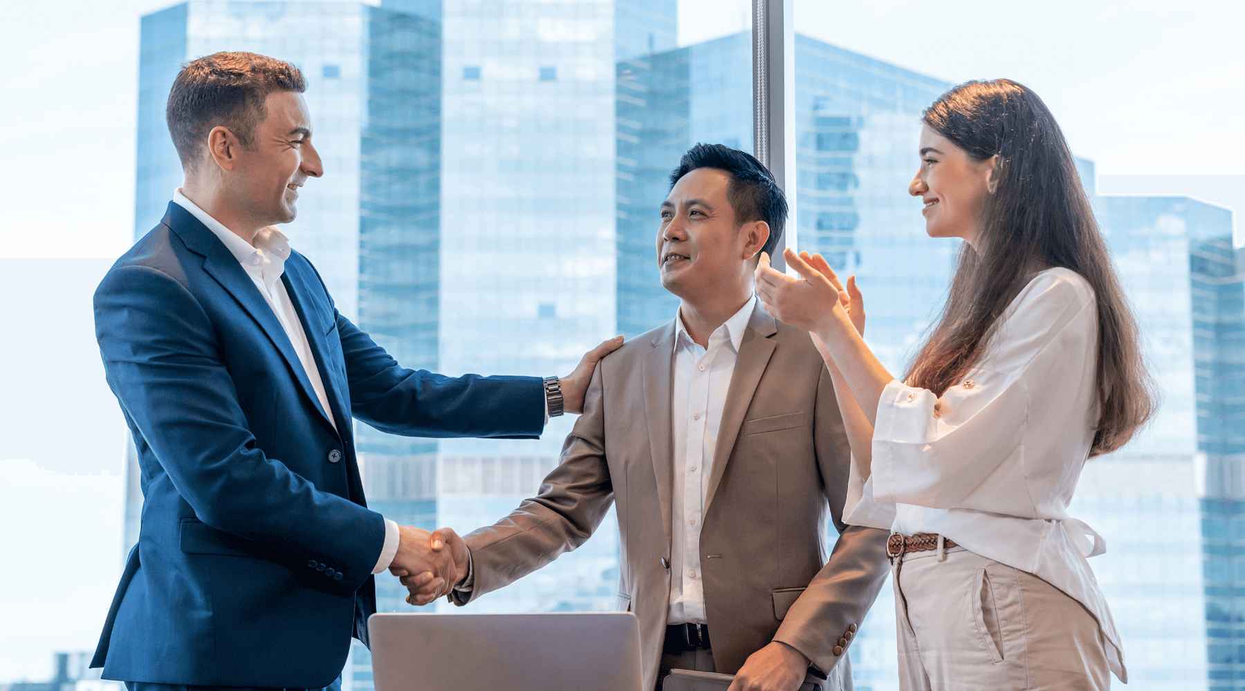 Three employees in a high-rise office are shaking hands and smiling.