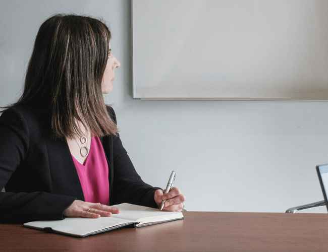 A woman wearing pink is writing in her notebook in a meeting.