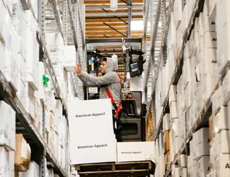 A man in a grey hoodie is operating a forklift accessing American Apparel boxes.