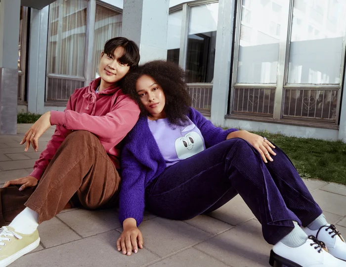 A woman wearing purple and man wearing pink are sitting on the floor leaning against each other.