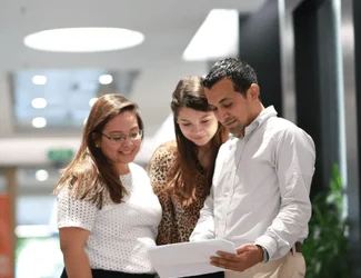 Three employees in Honduras are standing in the corporate office hallways looking at a paper.