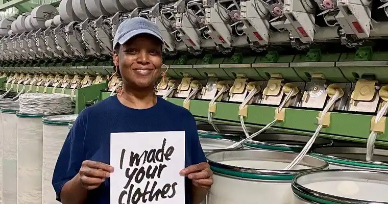 Jennifer sostiene un cartel que dice "Hice tu ropa" y sonríe a la cámara.