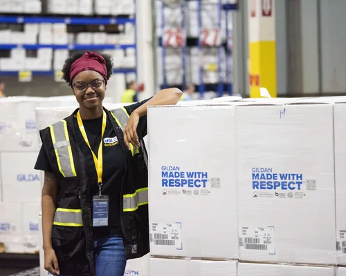 A female employee wearing a safety vest, standing next to stacked boxes with Made With Respect logo.