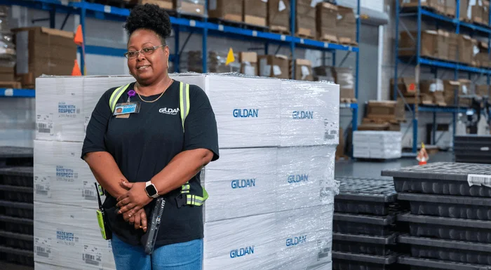 A woman in a black Gildan shirt is standing next to a stack of Gildan distribution boxes.