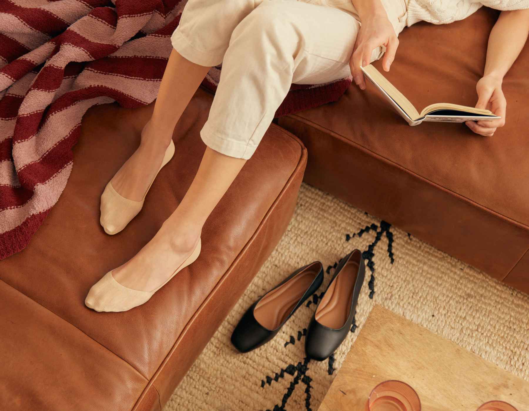 A woman sitting on a brown leather couch wearing Peds no show Peds socks and reading a book.