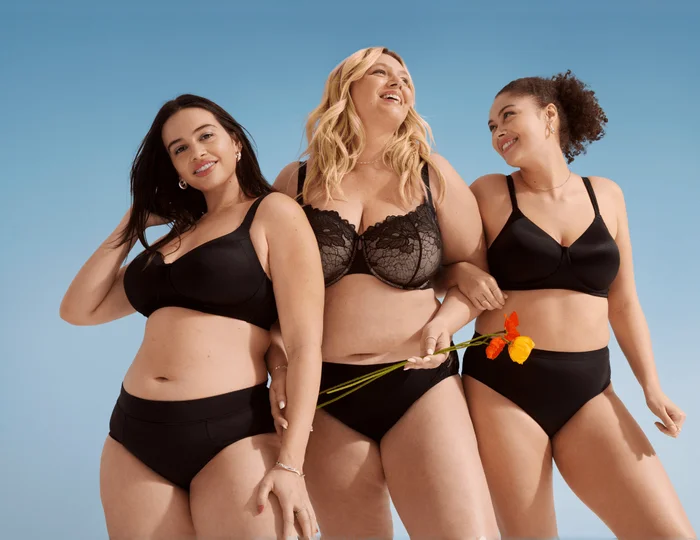 3 women wearing black innerwear are smiling together in front of a light blue background.