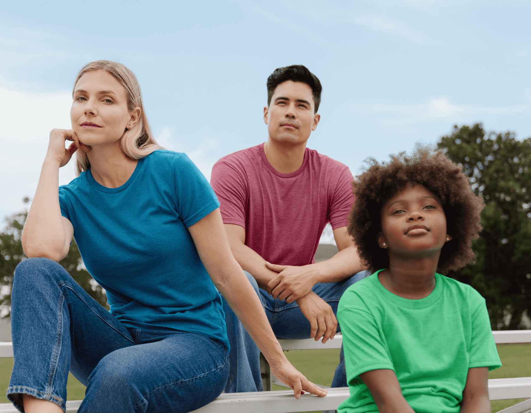 A woman, man, and boy wearing Gildan t-shirts are sitting outside on bleachers.