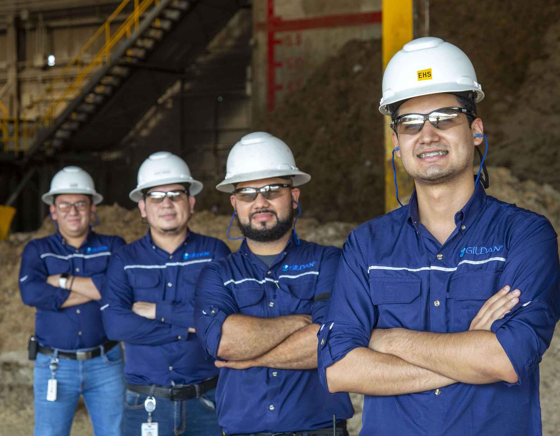 Cuatro empleados masculinos llevan camisetas Gildan y cascos sonriendo a la cámara con la Biomasa.