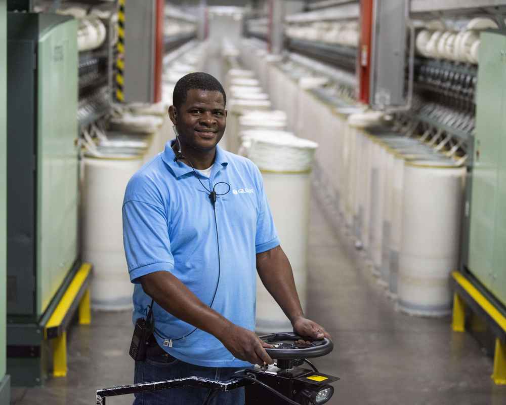 A man standing in the yarn spinning facility next to yarn cones smiling at the camera